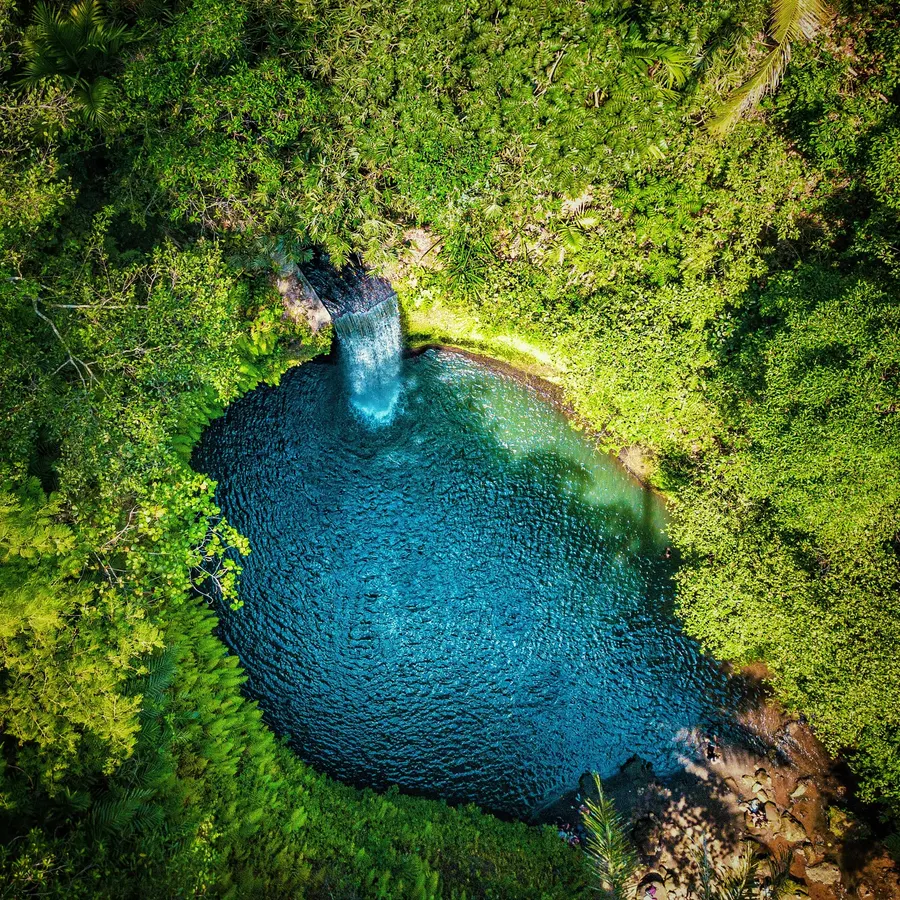 An aerial view of a small waterfall flowing into a bright blue pool, encircled by dense tropical greenery.