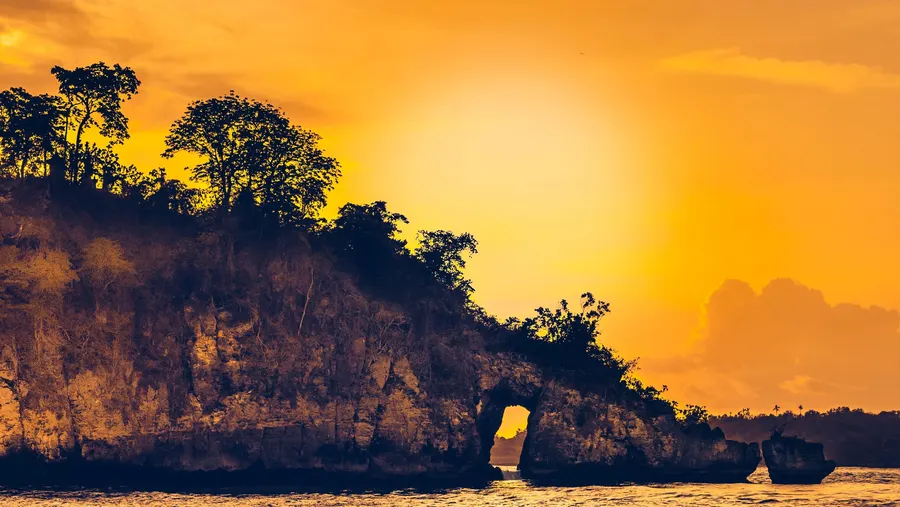 A rocky headland with a sea arch, silhouetted against an orange sky.