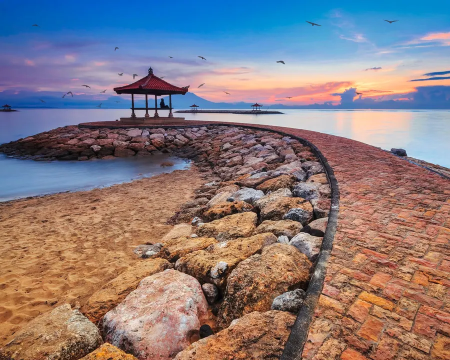 A stone pier curving out to a small pavilion under a pink dawn sky.