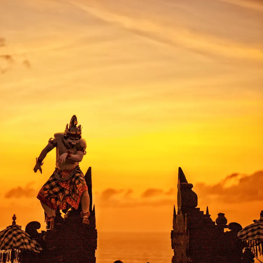 A Kecak dancer in an elaborate costume performing on a temple platform at Uluwatu Temple in Bali, silhouetted against a glowing sunset.