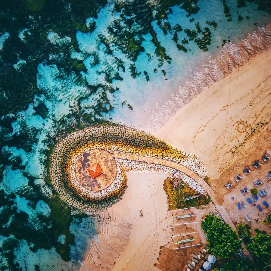 Aerial shot of Sanur Beach in Bali, showcasing a curved pier with a Balinese gazebo, surrounded by turquoise waters.