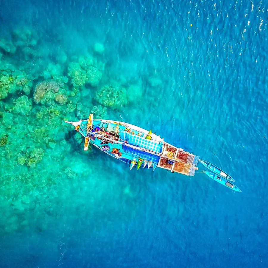 A colorful boat sailing on the clear blue waters near Komodo Island, with coral reefs visible beneath the surface.